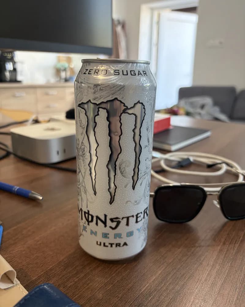 Fotka z mobilu: bílá plechovka Monster Energy Ultra na neuklizeném stole, kolem ní monitor, sluneční brýle, kabely a propiska