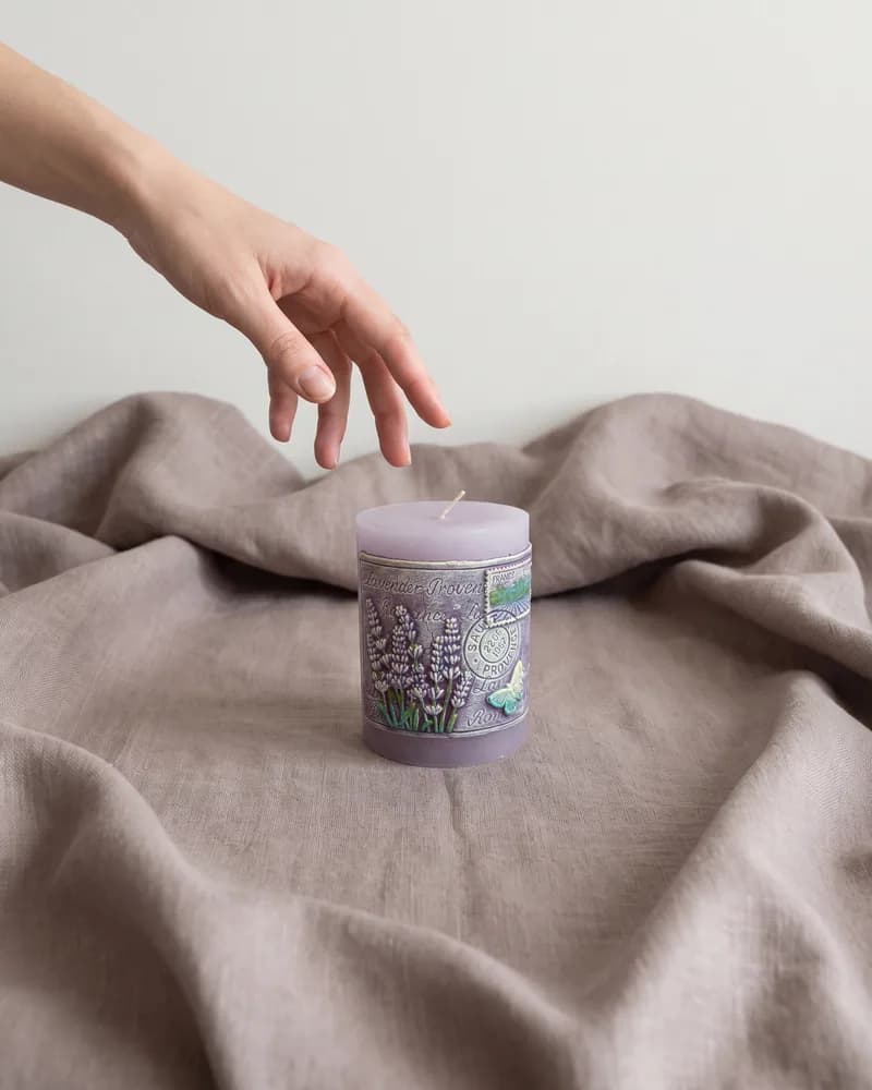 The lilac pillar candle on folds of taupe linen, a hand reaching down towards it, made with Nano Banana Pro.
