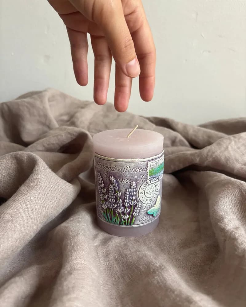 The lilac pillar candle on folds of taupe linen, a hand reaching down towards it, made with ProduktShot Lite.
