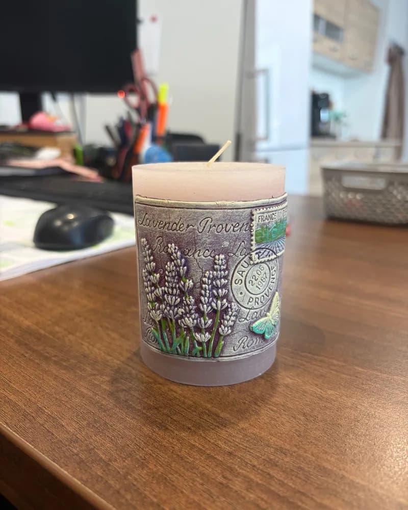 Ein Handyfoto einer fliederfarbenen Stumpenkerze auf einem Holzschreibtisch. Ihre geprägte Banderole zeigt Lavendel und einen Poststempel aus der Provence, im Hintergrund unscharf ein unaufgeräumtes Büro.